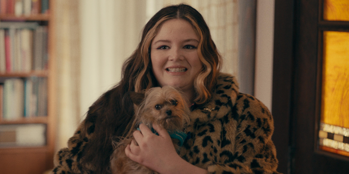 A still from "Cora Bora" where she is wearing a leopard jacket and holding a tiny dog with a awkward look on her face.