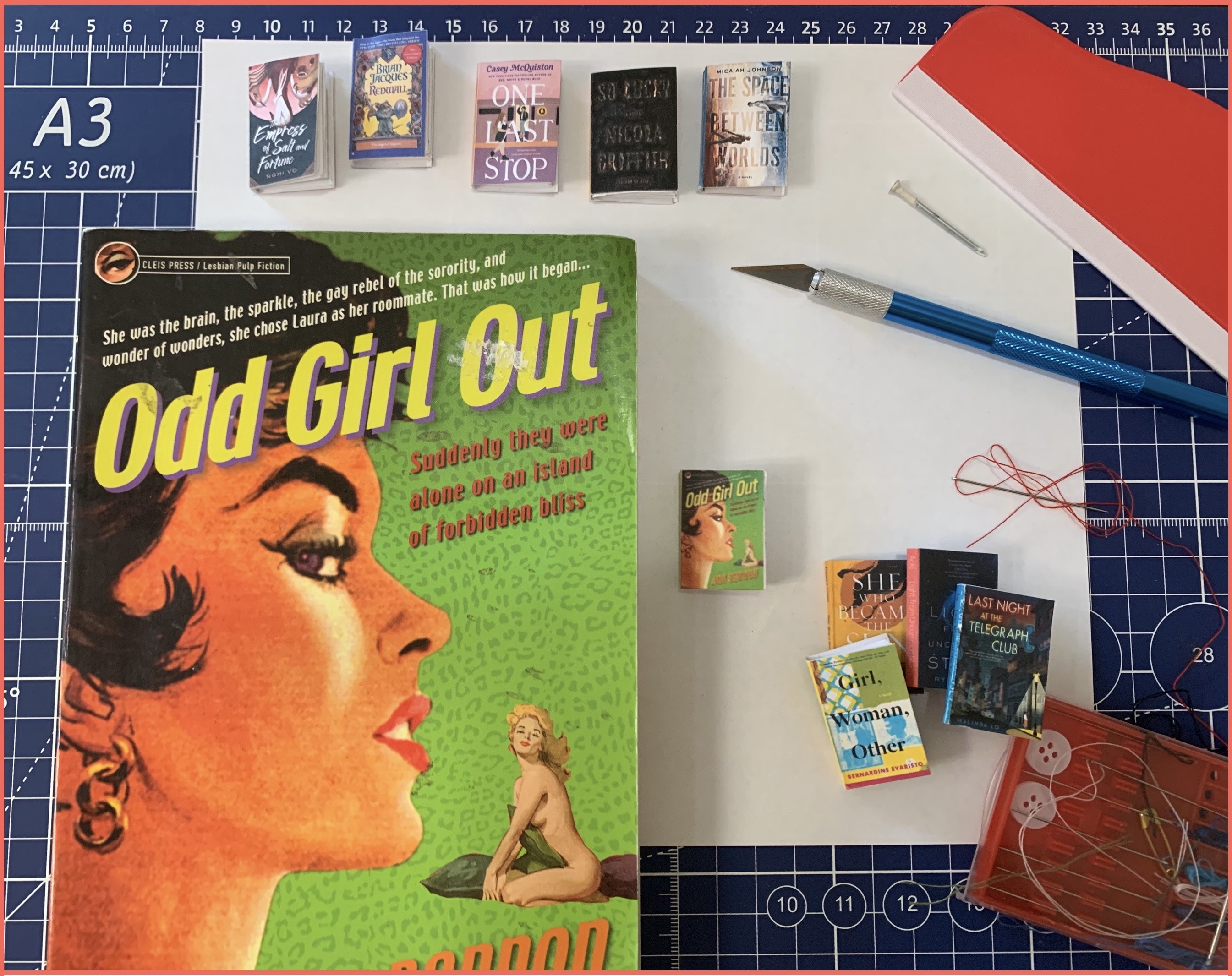 Assorted art supplies on a bright blue cutting mat.  Some tiny books and one normal sized book, a pulp fiction novel called Odd Girl Out.