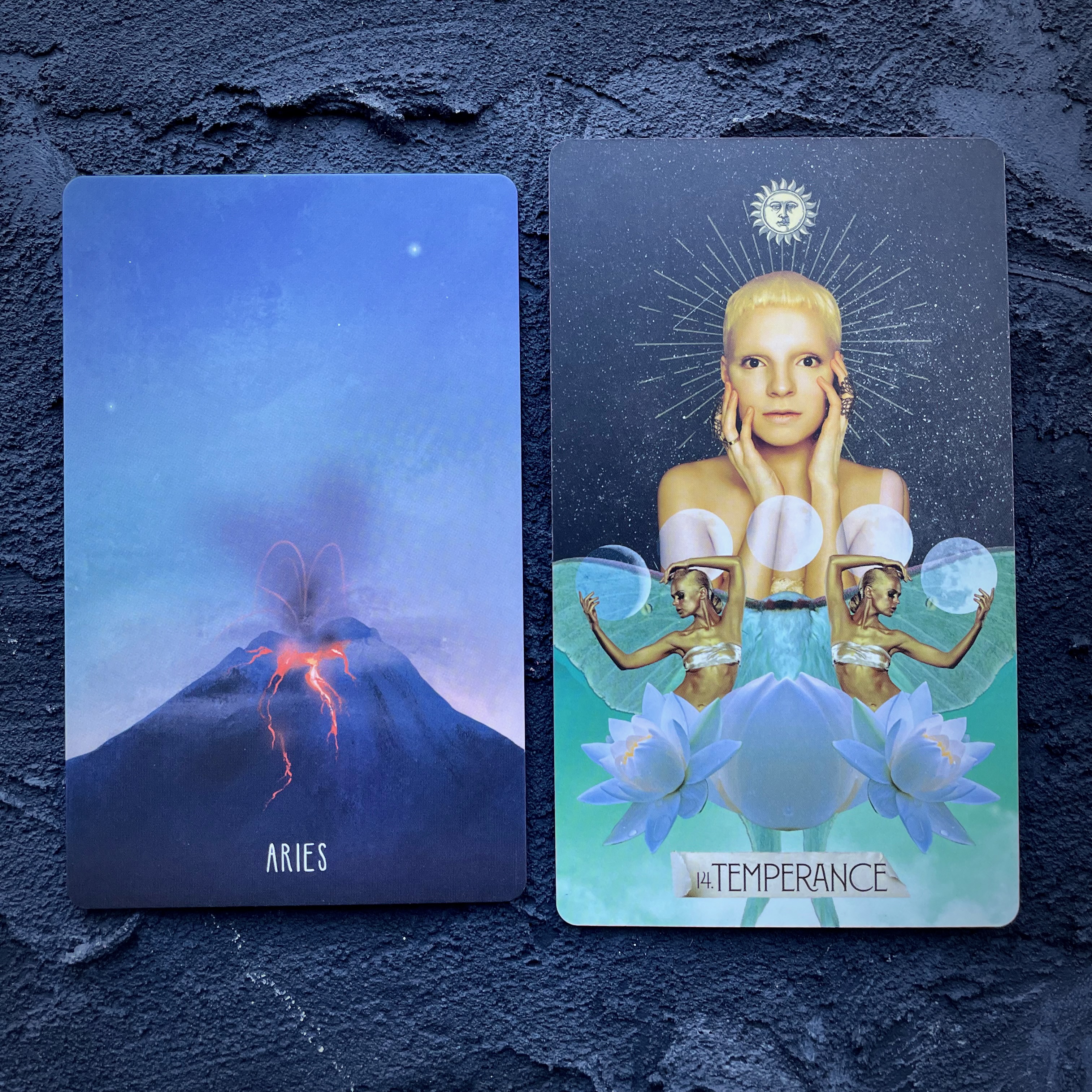 Two cards in front of a navy blue background, right to left: Aries (a volcano) and Temperance (a white person holding their face in their hands)