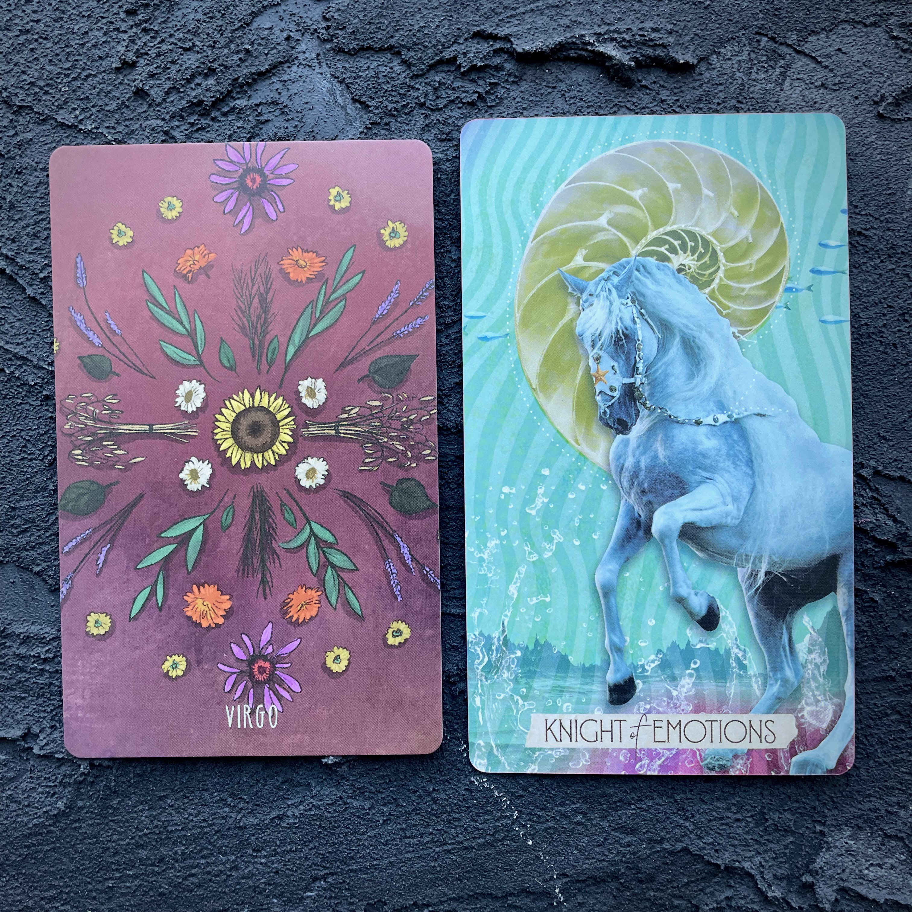 Two cards in front of a navy blue background, right to left: Virgo (spring blossoms) and Knight of Emotions (a unicorn)