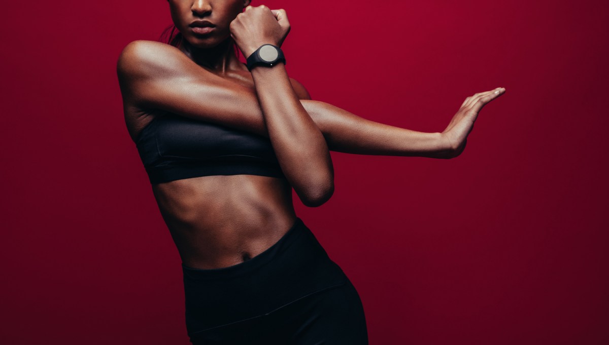 muscular woman stretching, wearing black workout gear and a watch in front of a red background
