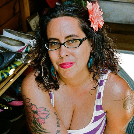 A woman with tattoos, glasses, and a flower behind her ear smiles confidently into the camera.