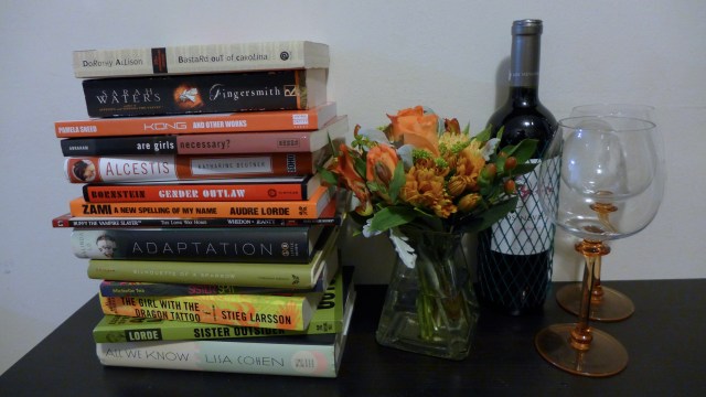 Stack of books next to a matching flower arrangement, a bottle of wine, and two wine glasses.
