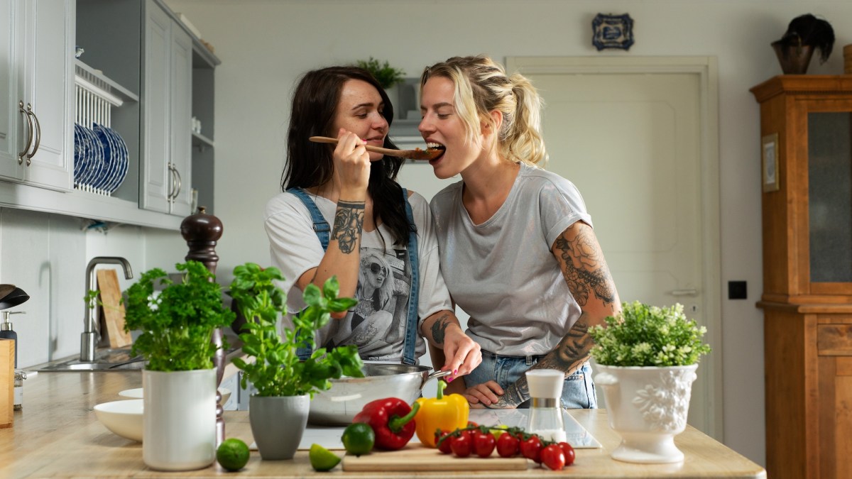 Tallinn, Harjumaa, Estonia - 06.17.2021: Beautiful Lesbian Couple Standing in a Modern and Sun Light Filled Kitchen Cooking Food Together As They Give Each Other a Taste With a Big Spoon.
