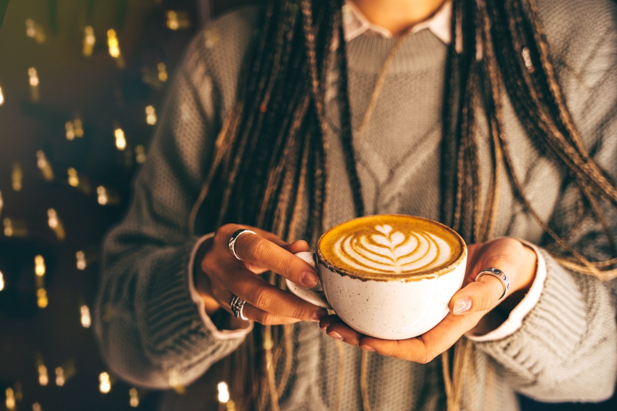 A woman with braided hair holding a cup of spiced Christmas coffee in her hands, close-up. Aromatic flat white or cappuccino with nice Rosetta latte art. Festive blurred and defocused garland lights on the background, space for text copy