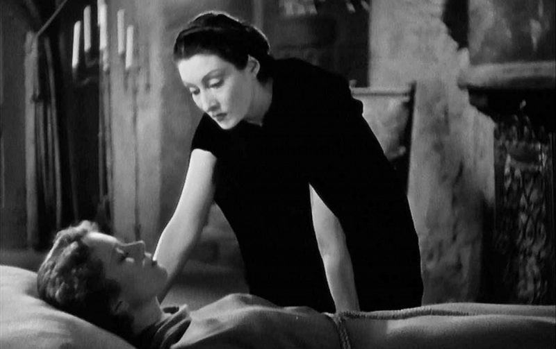 Queer horror to stream: Dracula's Daughter. Gloria Holden as Countess Marya Zaleska is wearing all black and hovering over a sleeping young woman.