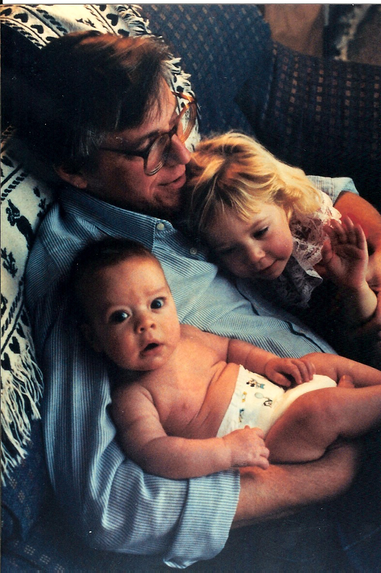 a very young rachel and her sibling (a baby) are snuggling with their dad on a couch