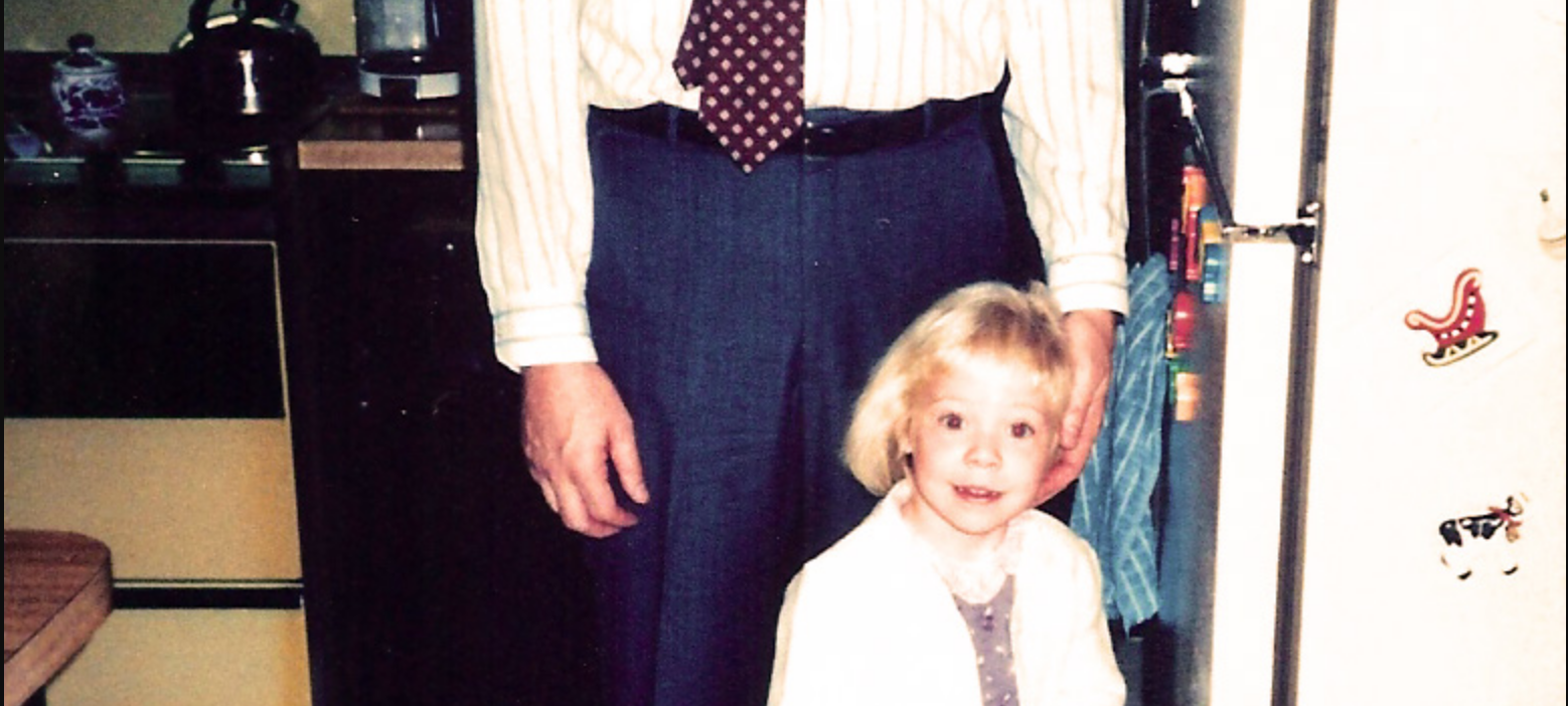 a very small Rachel, with blonde hair, is seen standing in front of an adult man who is wearing slacks, a white shirt and a tie. you cannot see the man's face due to the cut of the photo