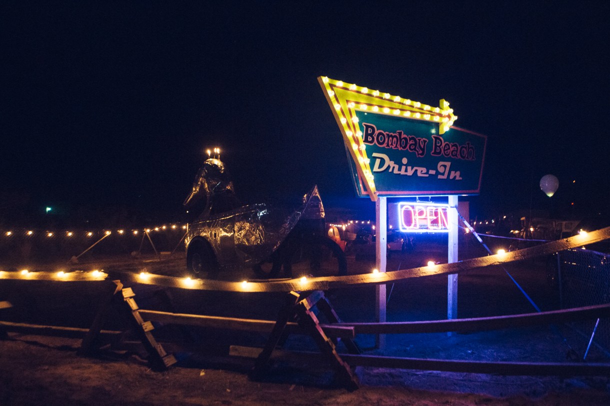 A drive-in installation filled with destroyed cars and other vehicles screening films like Fellini’s 8 1/2