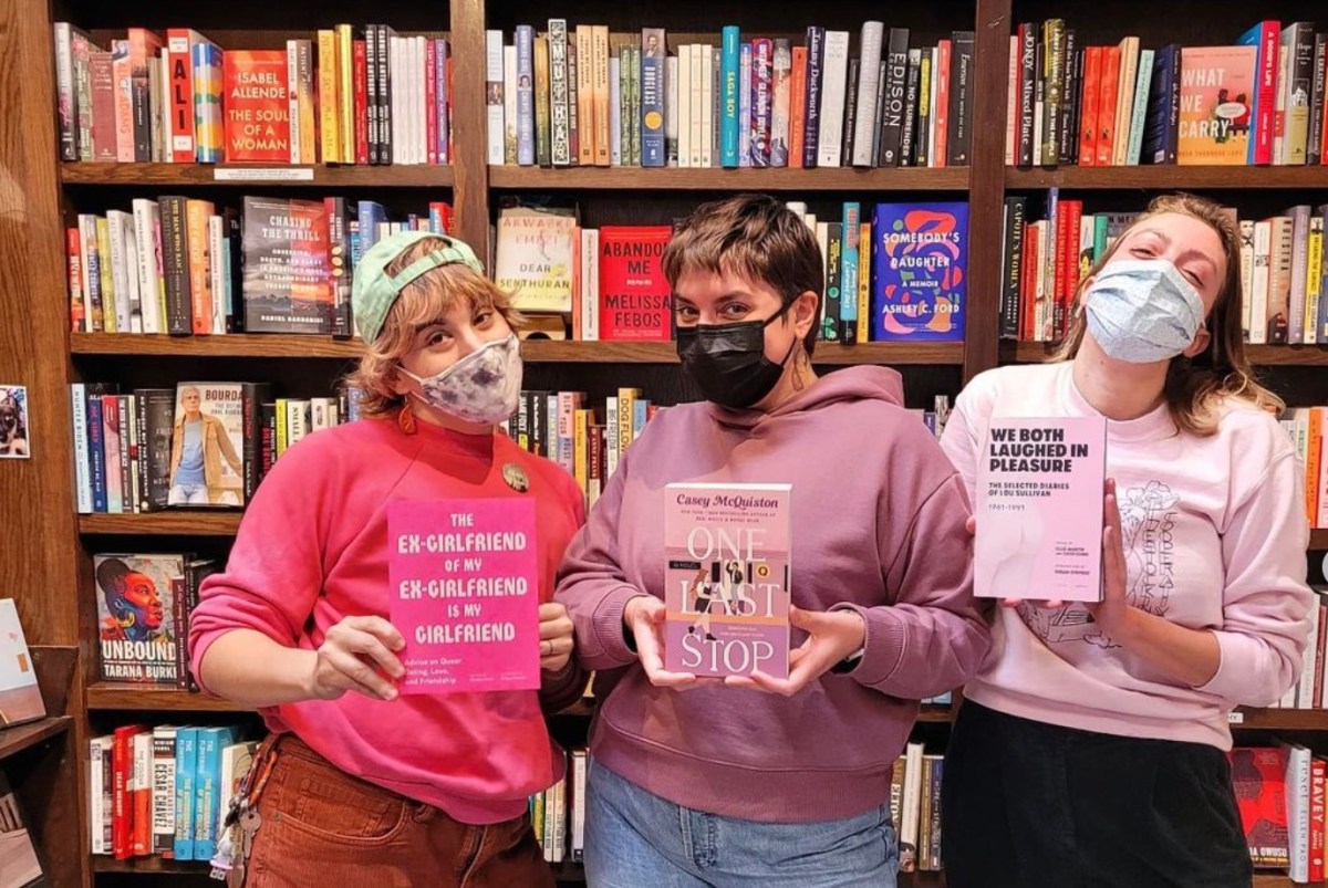 three humans holding up queer books at a Room of Ones Own Bookstore