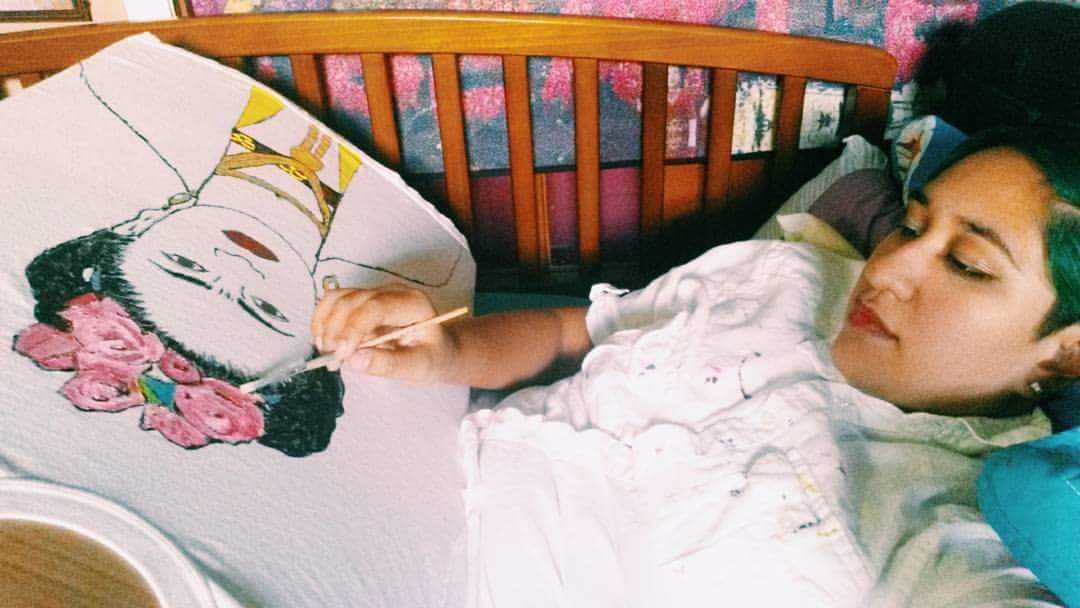 Annie, a young Latinx woman with green hair, lying in bed painting a portrait of the artist Frida Kahlo.