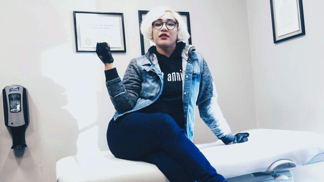 Annie, a young Latinx woman with blonde hair, circular black glasses, and red lips in this photo, poses on the examination table in a doctor's office. One had is up in the air in a fist.