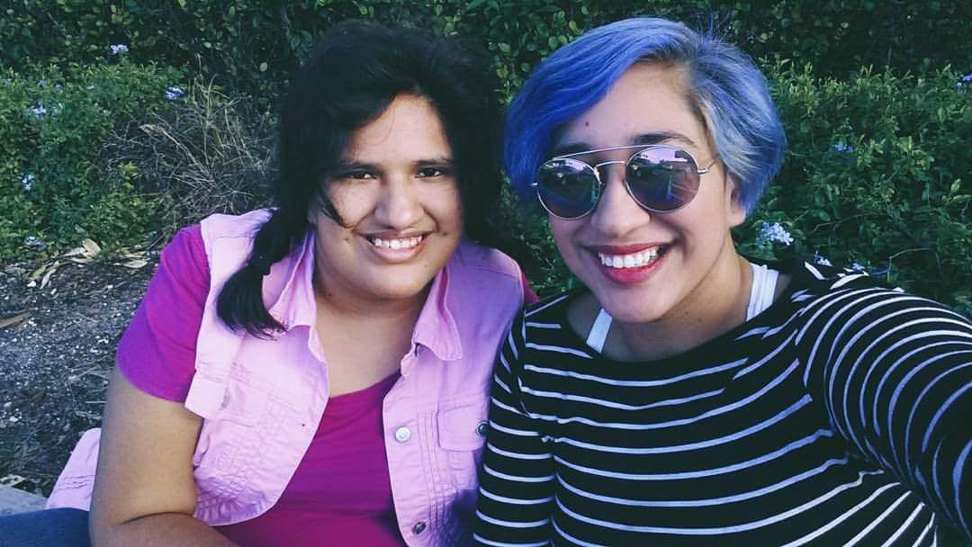 Emily and Annie, two Latinx sisters in their twenties, sit side by side smiling at the camera. Emily has dark hair and is wearing a dark pink shirt with a lighter pink vest over it. Annie has blue hair and is wearing a black shirt with white stripes, as well as sunglasses.