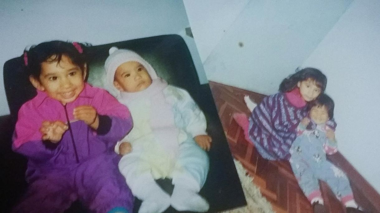Two photos side by side. On the left, sisters Emily and Annie are very young. Emily is in a white onesie and Annie is in a pink and purple jumpsuit. In the right-hand photo, both siblings are slightly older but still children, with Annie putting her arms around Emily to hug her from behind. Annie wears purple and pink, Emily is in blue denim overalls.