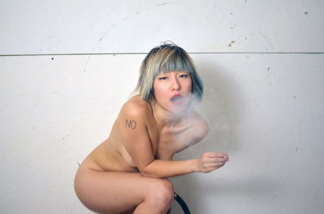 styrxfoam sitting naked on a stool against a white wall, exhaling smoke