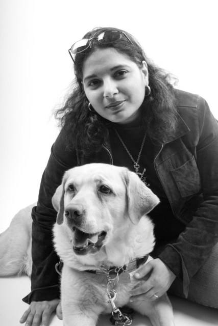 An Arab-American woman with dark hair, a black jacket, and sunglasses perched on her forehead looks into the camera. She is sitting with her arm around a large dog with light-colored fur. The photo is black and white.