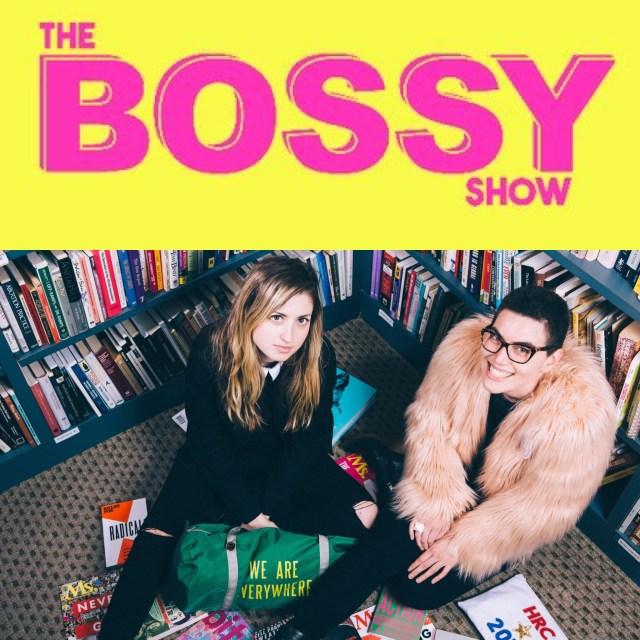 The top part of the photo is the logo for The Bossy Show: the show title in all caps, bright pink font on a bright yellow background. Below, a white woman in a black shirt and black pants with shoulder-length blonde hair and a Latina woman in a pink fur coat with buzzed dark hair and glasses sit side by side on a library floor, surrounded by bookshelves and smiling up at the camera. Feminist magazines are scattered on the floor around them, along with a green duffel bag that reads "We are everywhere."