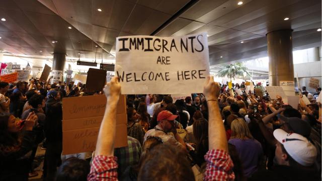 A woman holds up a black and white sign in a large crowd at an airport protest that reads "Immigrants are welcome here."