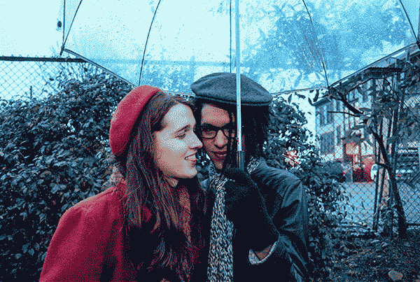 Moving image GIF of Yael (a white woman in her twenties with long brown hair and wearing a red coat and beret) kissing her girlfriend Jarreau (a black woman in a black leather jacket with a gray scarf and beret) on the cheek.