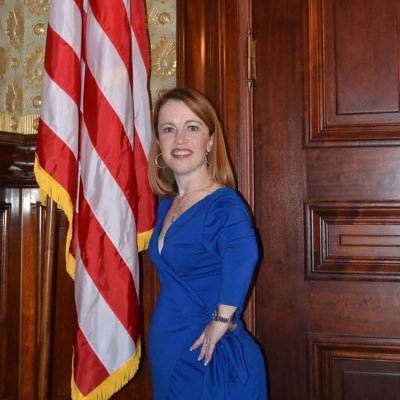 A white woman with long red hair and red lips stands next to an American flag in a room with a dark wooden door. She is wearing a blue dress and has her hand on her hip.