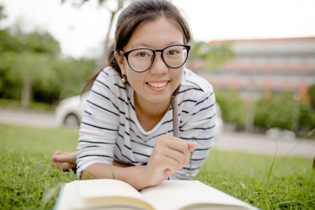 girl with oversized glasses reading a book outside on the grass