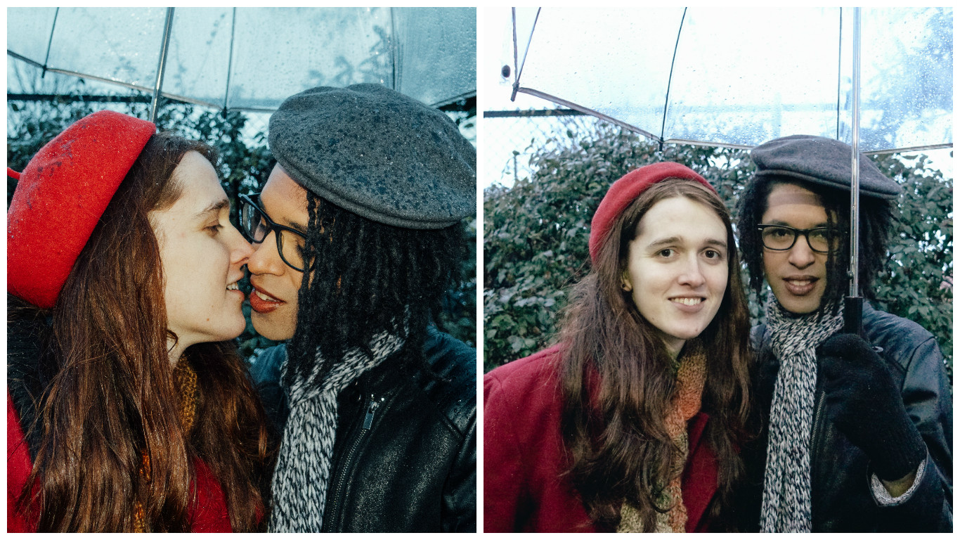 Two photos side by side: on the left, a white woman in her twenties with long brown hair and wearing a red coat and beret and a black woman in a black leather jacket with a gray scarf and beret lean toward each other in a kiss. On the right, the two of them face the camera, smiling. They are under an umbrella in both photos.