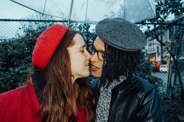 Yael (a white woman in her twenties with long brown hear and wearing a red coat and beret) and her girlfriend Jarreau (a Black woman in a black leather jacket and a gray scarf and beret) lean in for a kiss under a clear umbrella