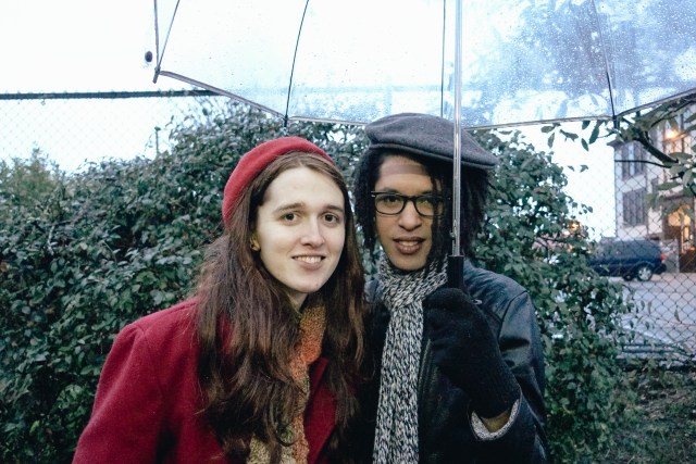 Yael (a white woman in her twenties with long brown hear and wearing a red coat and beret) and her girlfriend Jarreau (a Black woman in a black leather jacket and a gray scarf and beret) smile at the camera under a clear umbrella