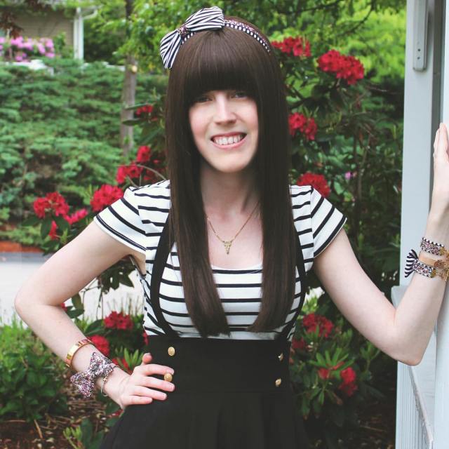 A white nonbinary person with long brown hair and blunt bangs stands against a backdrop of a hedge with red flowers on it. She is wearing a black and white striped dress and has her hand on her hip.