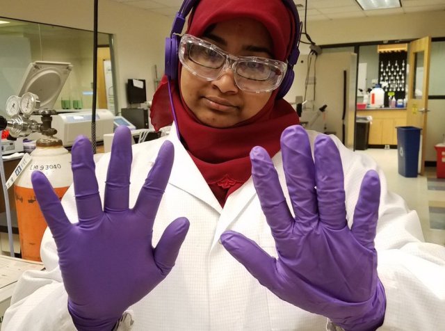 A person in a hijab and lab coat with purple gloves and safety goggles.