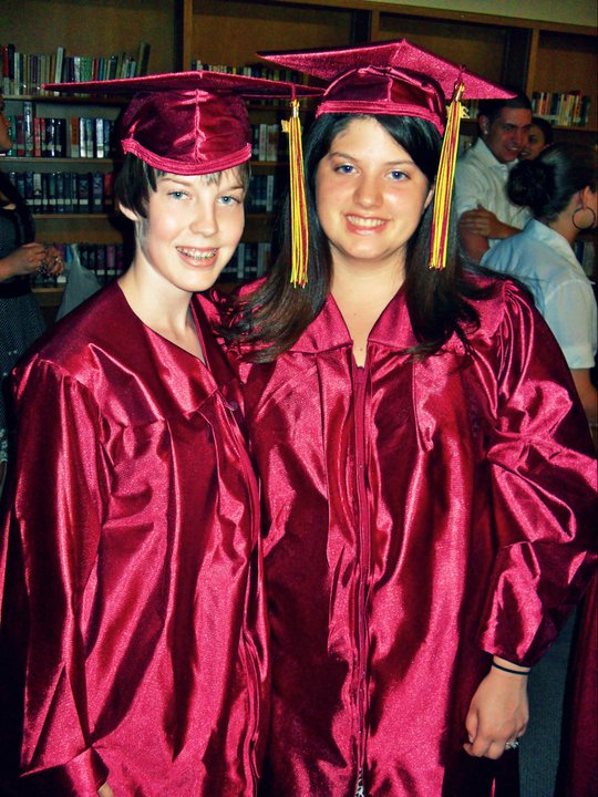 Two people smile into the camera wearing burgundy graduation caps and gowns with gold tassels.