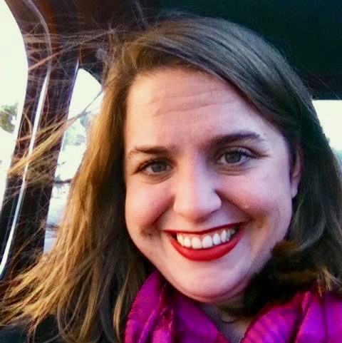A young white woman with long brown hair and wearing red lipstick and a pink scarf smiles widely into the camera
