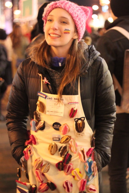 A person wearing a pink "pussy hat" and an apron with lots of buttons and sculpted vulvas pinned to it
