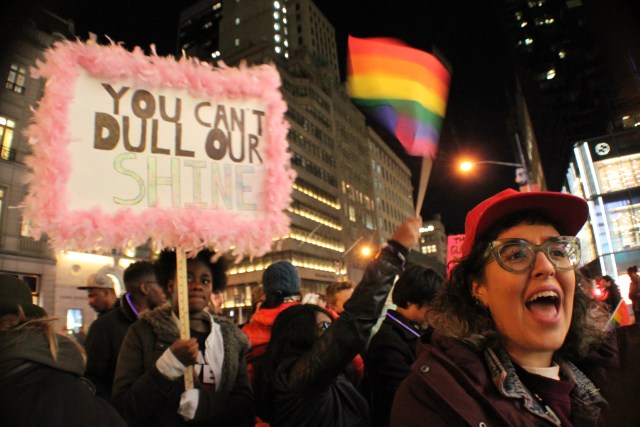 A group of protesters, one waving a rainbow flag, one holding a sign that reads YOU CAN'T DULL OUR SHINE, and one person shouting or chanting