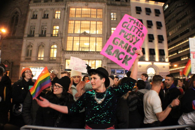 A group of protesters, with one person in the foreground wearing a glitter shirt and holding a sign that reads THIS GLITTER PUSSY GRABS BACK