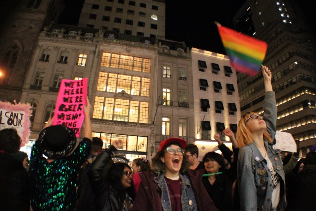 A group of protesters in motion, waving a rainbox flag and a sign that reads WE'RE HERE WE'RE QUEER WE'RE NOT GOING ANYWHERE