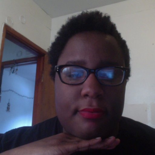 A black woman with short curly hair, red lipstick, and thick black glasses looks into the camera with an open doorway in the background