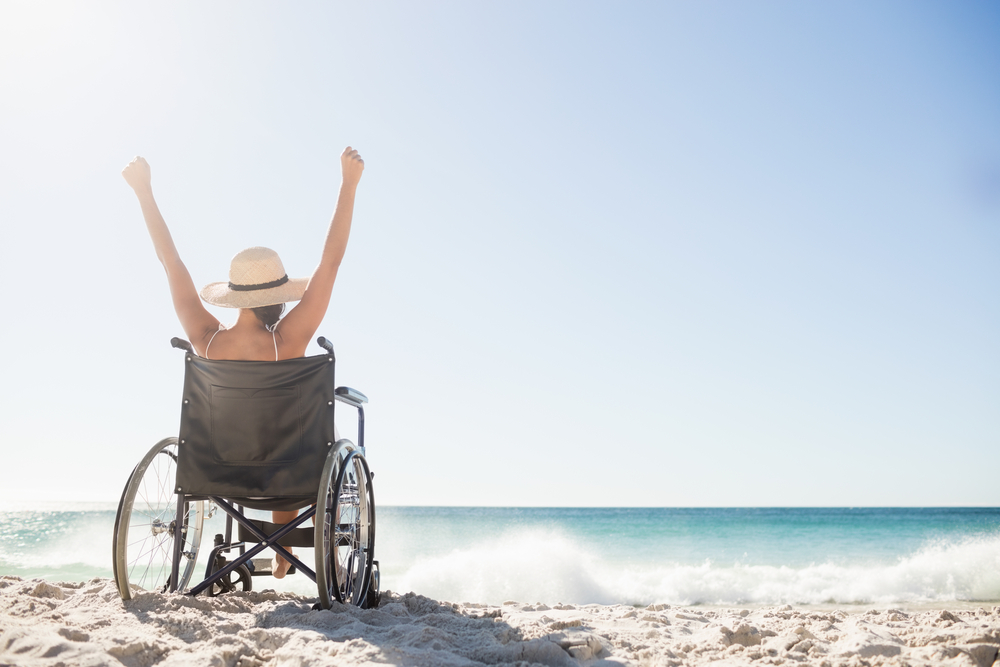 View from behind of a woman raising her arms over her head while sitting in a manual wheelchair on the beach. She is wearing a straw hat with a wide brim, and you can see the ocean in the background.