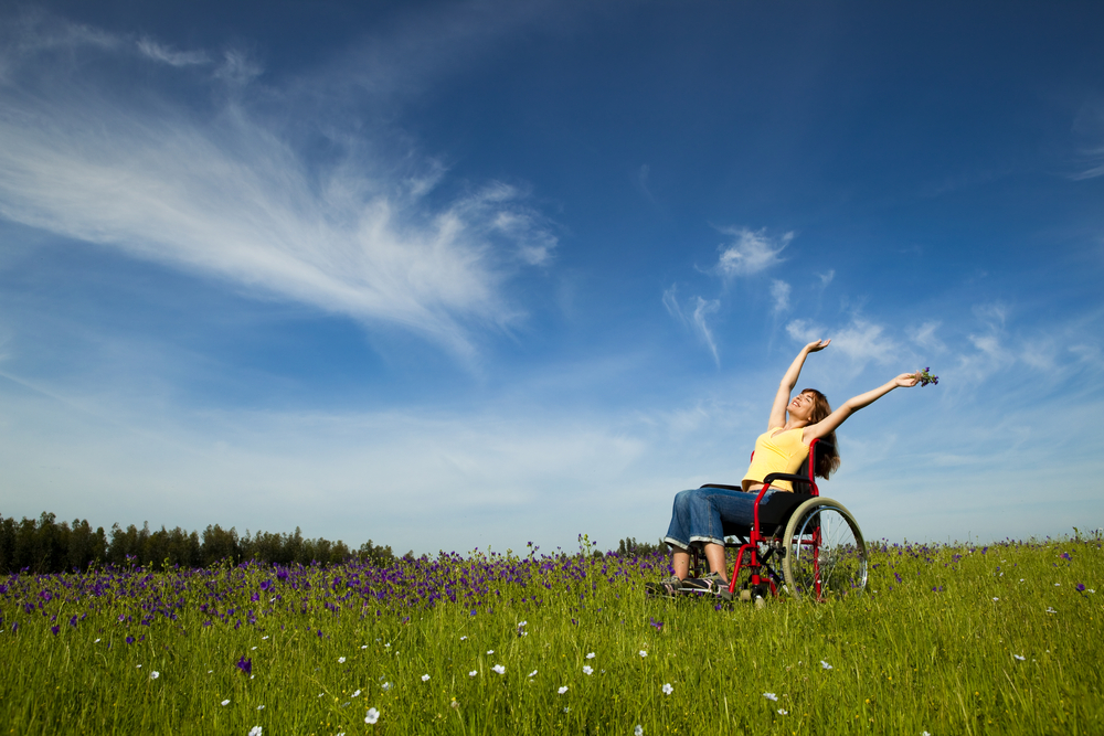 A woman in blue jean capris and a yellow sleeveless top raises her arms over her head while sitting in a red manual wheelchair in the middle of a meadow.