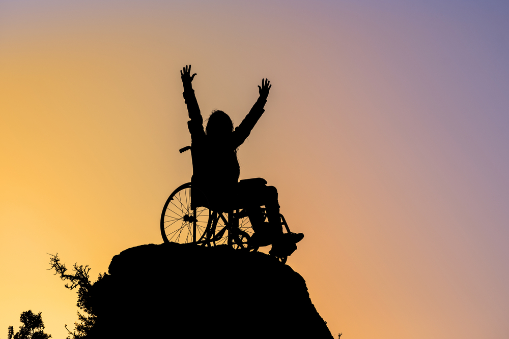 In silhouette against a sunset background, a person in a manual wheelchair raises their arms above their head on a mountaintop.