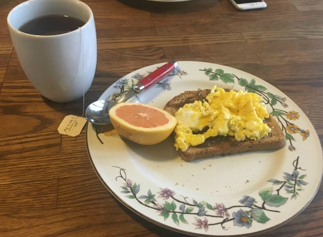 A plate of food showing eggs on toast with a small grapefruit slice next to it. There is a white mug full of tea in the background.