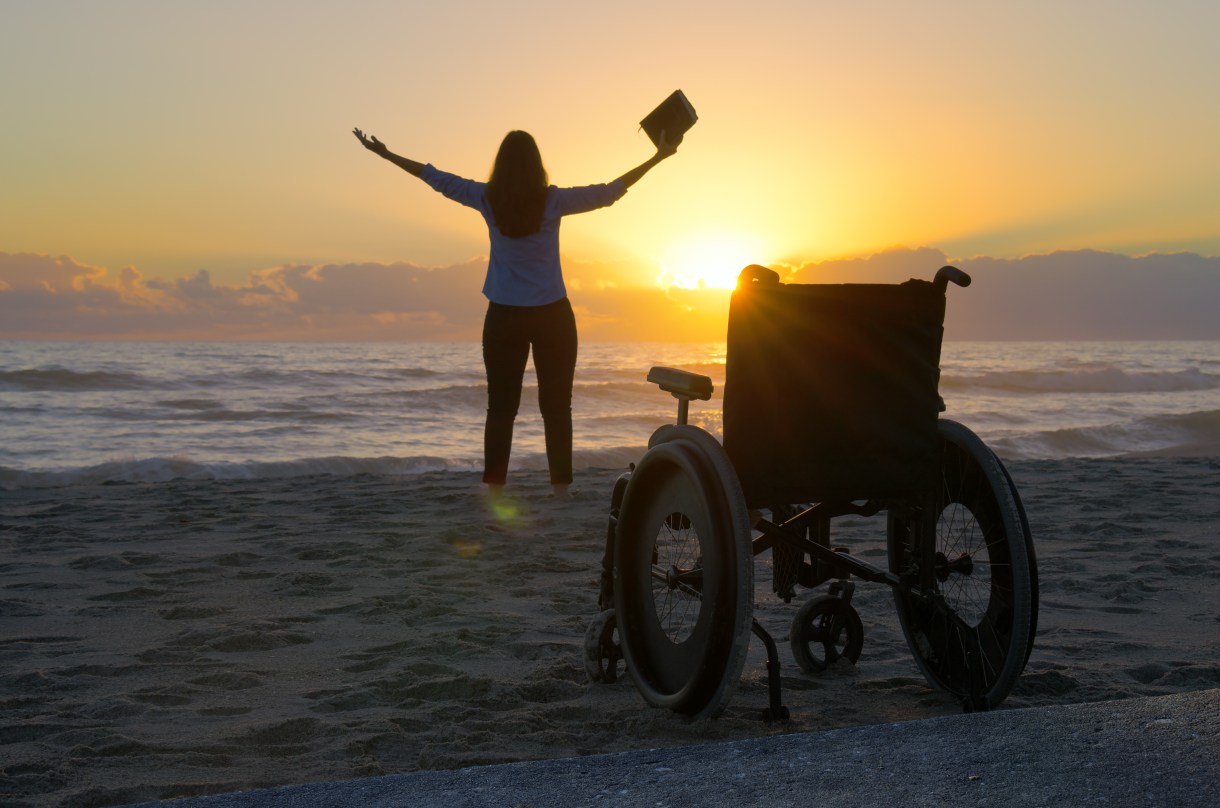 In silhouette against a sunset background, a woman raises her arms towards the sky and holds a book in one hand. A manual wheelchair sits unoccupied toward the back of the frame.