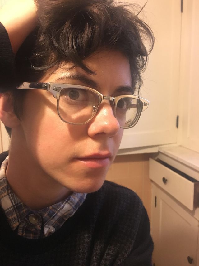 A white woman with short brown hair and glasses fixes her hair by running her fingers through it. She is wearing a blue and white plaid shirt underneath a black houndstooth sweater.