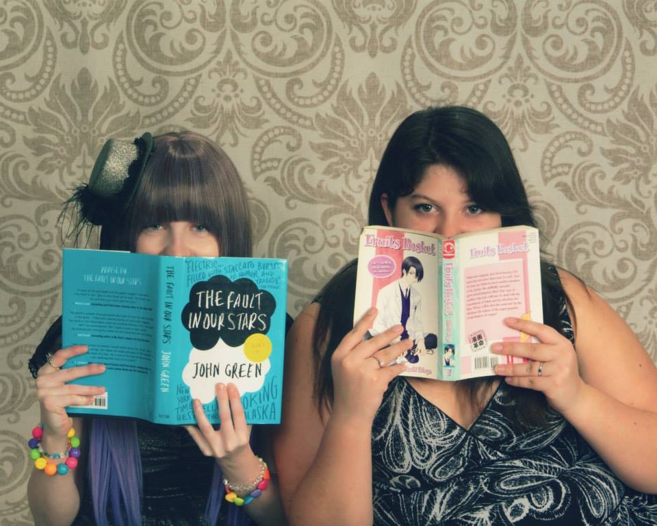 Two people jokingly hide their faces behind book covers. The person on the left has long brown hair cut into severe bangs and is wearing a small hat tilted off the right side of their head. They're behind the book cover of The Fault in Our Stars. The one on the right has darker, longer hair and is wearing a black and white top. They're behind the cover of a book in the Fruits Basket anime series.