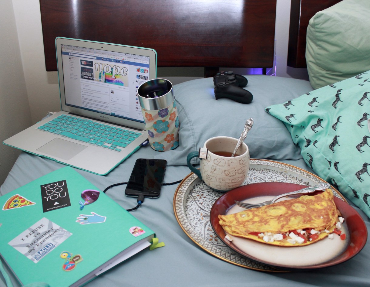 Photo of a bed. There is a tray with tea and an omelette, a passion planner, colorful pens in a tea tin, a laptop, a Playstation controller, and a plugged in phone. Everything is teal.