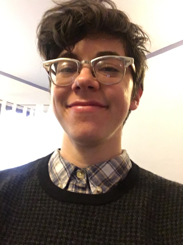 A white woman with short brown hair and glasses wearing a blue and white plaid shirt under a black houndstooth sweater smiles close-lipped at the camera.