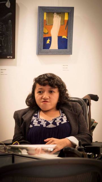 A young Asian woman with shoulder-length hair and wearing a black shirt with white trim sits in her wheelchair looking at the camera. There is a painting of a foot against a blue and orange background on the wall behind her.