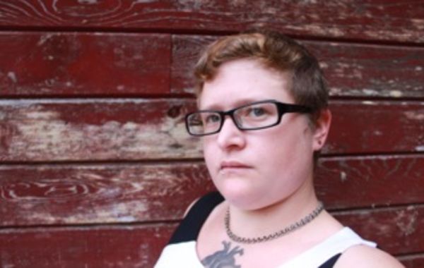 A white person with curly brown/blonde hair and square-framed black glasses stands against a brick wall background