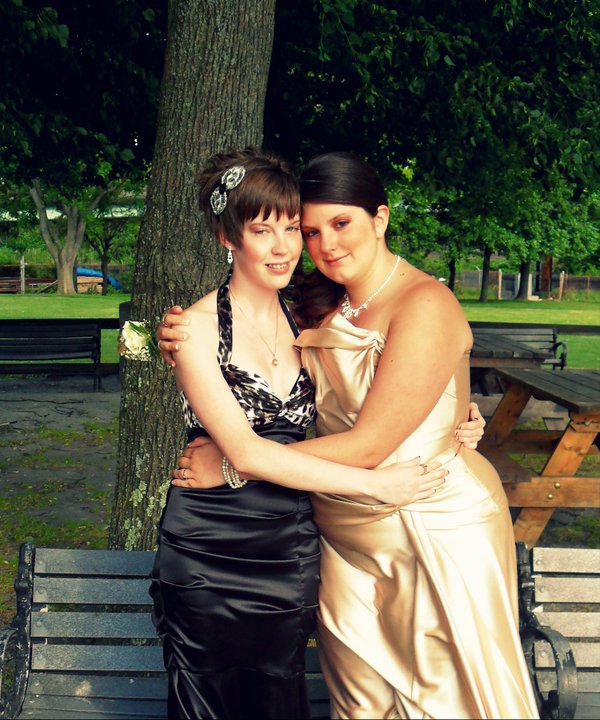 Two people hold each other around the waist in fancy prom dresses. The one on the left is in a black dress, while the person on the right is in gold.