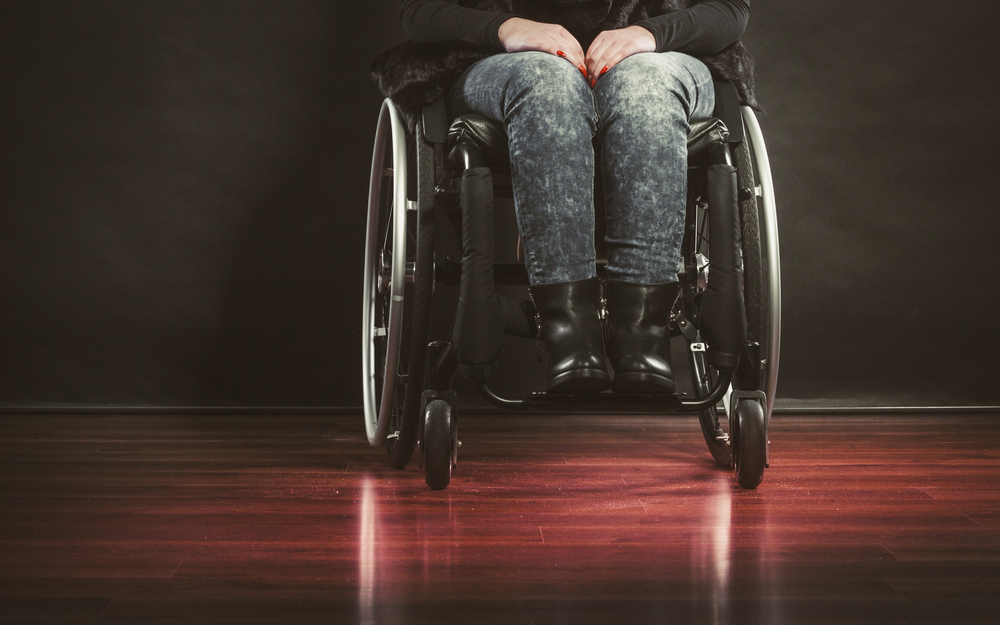 Front-facing view of a woman's legs in a manual wheelchair. She is wearing black stonewashed jeans and black boots, and is in a room with a dark wooden floor. You can only see her legs in the chair, no head or torso.